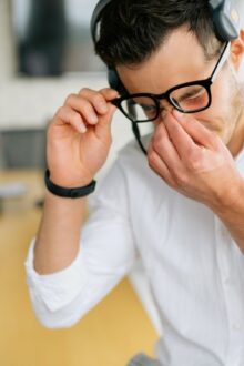 A man sits on a desk in the office rubbing his face, appearing to have a headache